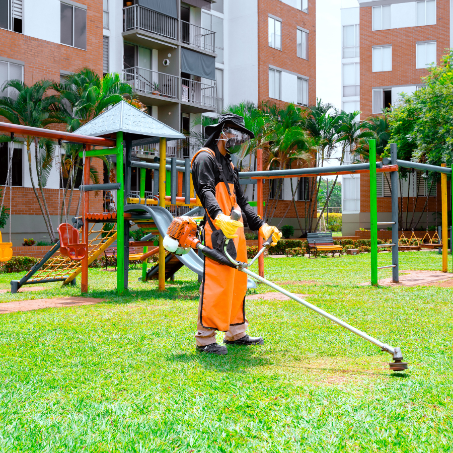 Operario de jardinería con protección facial y equipo de aspersión listo para el control fitosanitario en el área de juegos de un conjunto residencial.